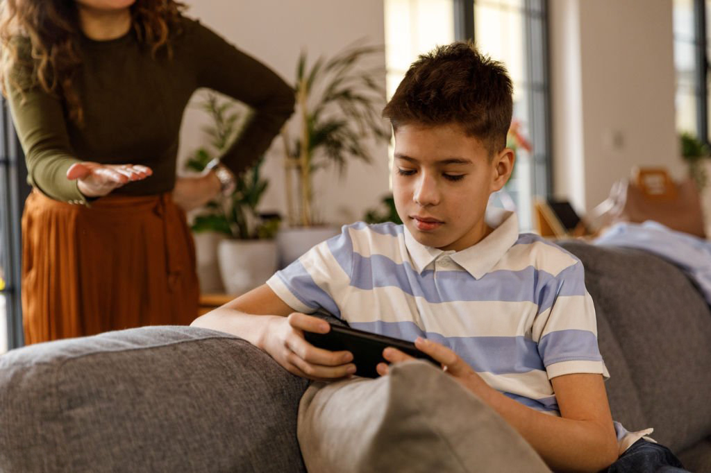 Photo en mise au point sélective d’un adolescent allongé sur le canapé, passant son temps à jouer à des jeux vidéo sur son smartphone. Sa mère, en colère, se tient derrière lui, la main sur la hanche, observant ce qu’il fait et lui demandant de lui donner le téléphone.
